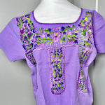 Load image into Gallery viewer, A lavender purple short-sleeve blouse with colorful floral embroidery on the chest and front panels, displayed on a white mannequin torso in front of a gray door.
