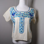 Load image into Gallery viewer, An off-white short-sleeve blouse with blue and green floral embroidery on the chest, displayed on a white mannequin torso against a gray door background.
